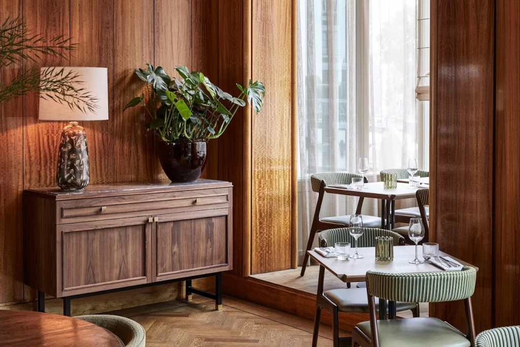 Элегантный интерьер ресторана с деревянными панелями на стенах, зелеными стульями с подушками и деревянным буфетом с лампой и большим комнатным растением. В ресторане уютная и изысканная атмосфера, аккуратно сервированные столы и большие окна, пропускающие естественный свет.